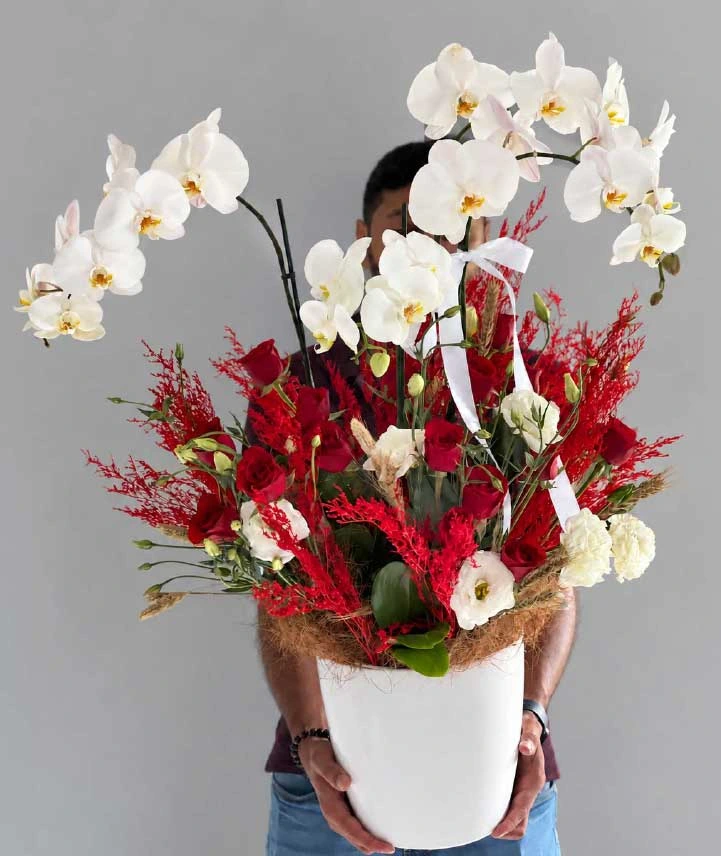 arreglo-con-orquídeas-blancas-y-rosas-pasión-imperial Arreglo floral premium de orquídeas Phalaenopsis blancas y rosas rojas en elegante recipiente blanco con detalles botánicos rojos.