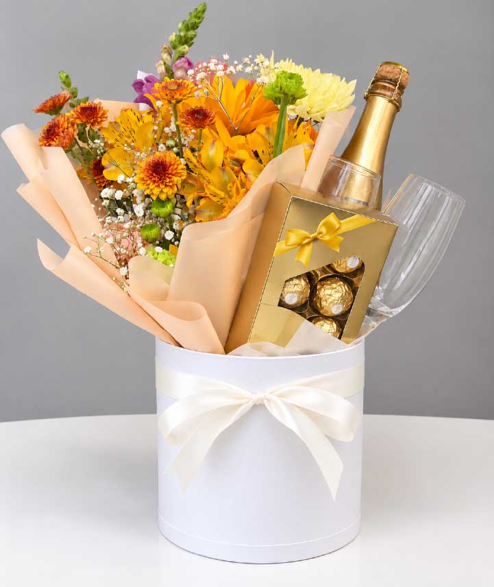 Caja floral con flores naranjas y amarillas, vino espumoso, chocolates premium y copas en elegante caja blanca.